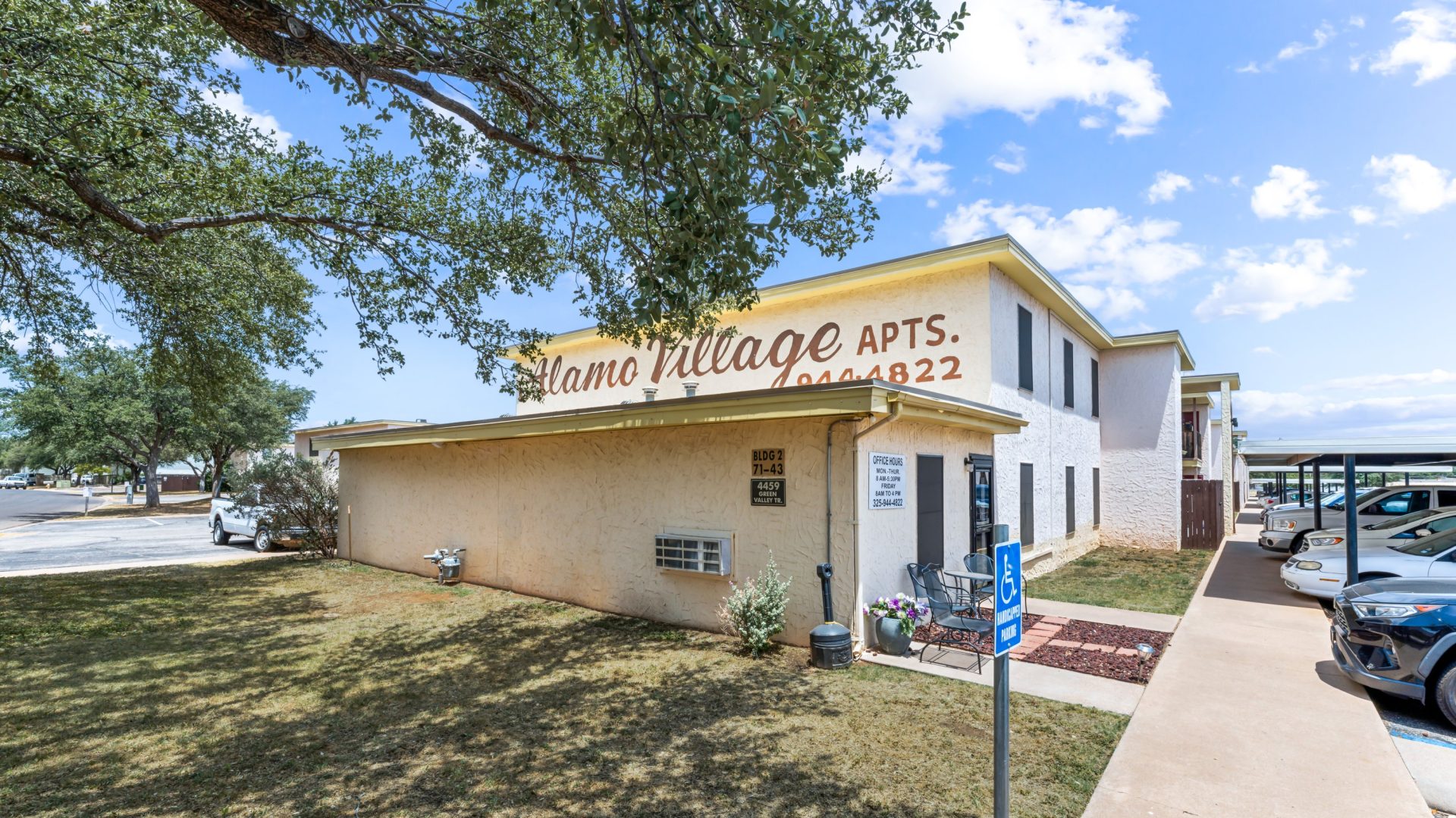 the outside of a motel with a parking lot and trees at The Alamo Village Apartments